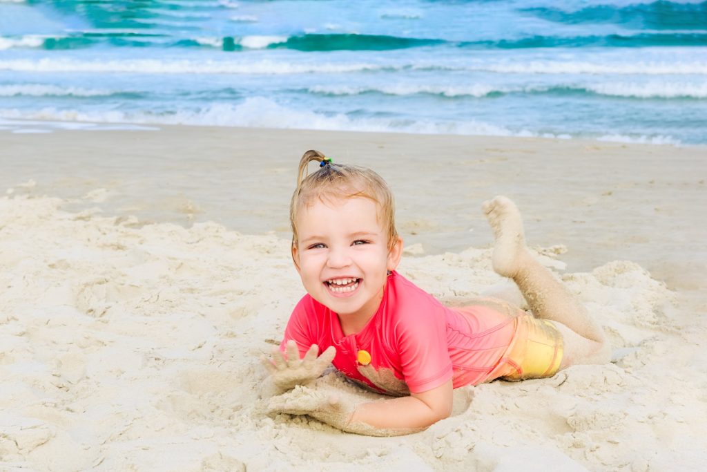 Laughing toddler girl in sun protecting swimming suit lying on sand on the beach. Family vacation